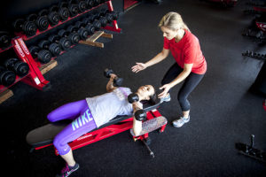 A woman in red supports a woman in purple as she lifts weights on a bench